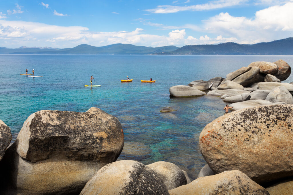Kayakers and paddleboarders paddling on the turquoise crystal-clear water of Sand Harbor in Lake Tahoe with giant boulders in the foreground and mountains in the background